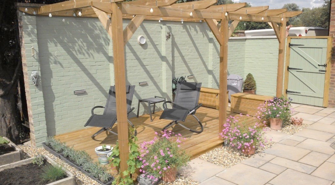 A medium-sized lean-to pergola over some decking, strung with decorative lights and surrounded by assorted potted flowers.