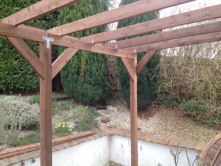 A pergola constructed in front of a small treeline, stained with a walnut-brown wood stain
